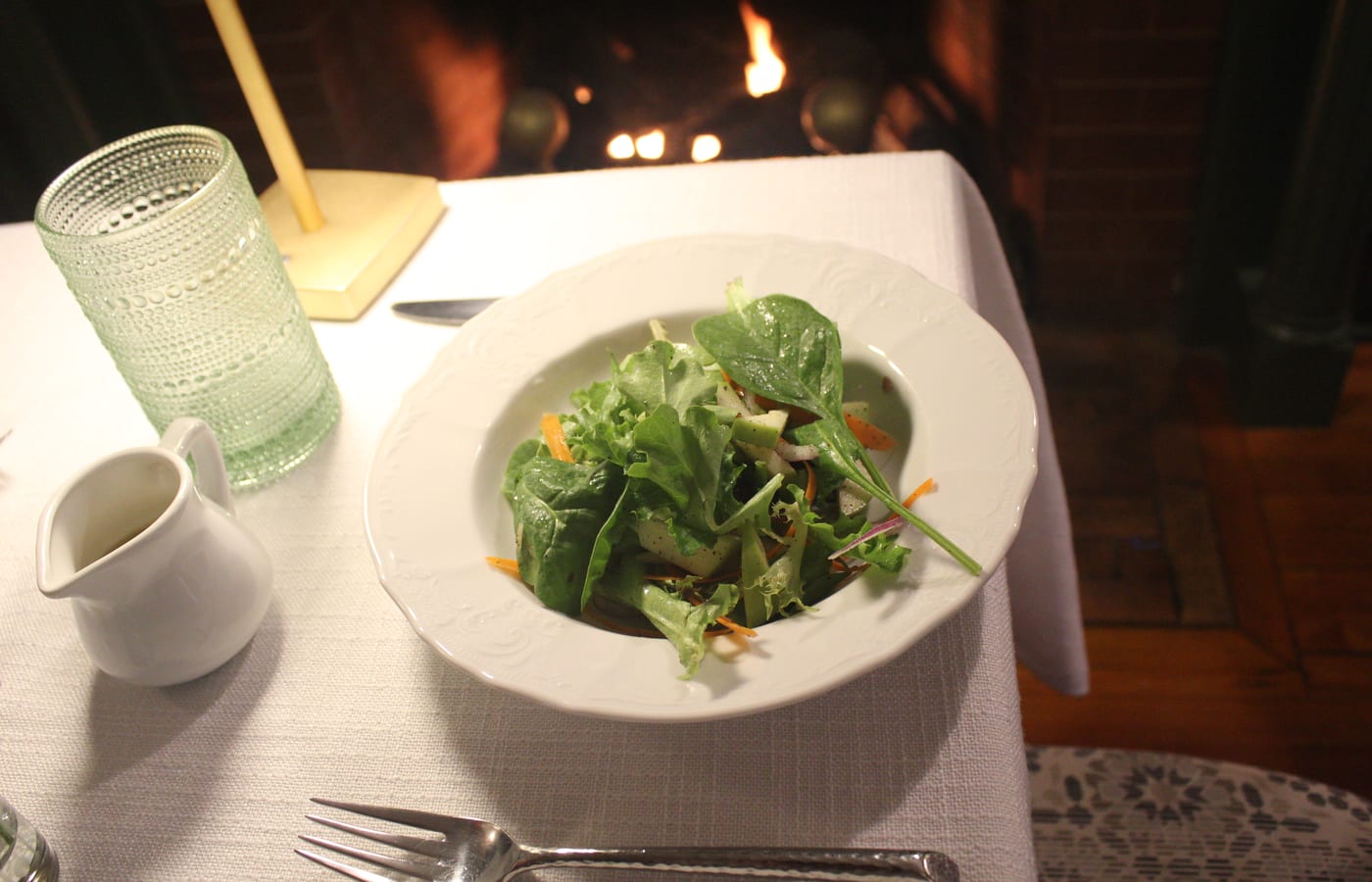 A salad course on a linen-set table with a fire burning behind